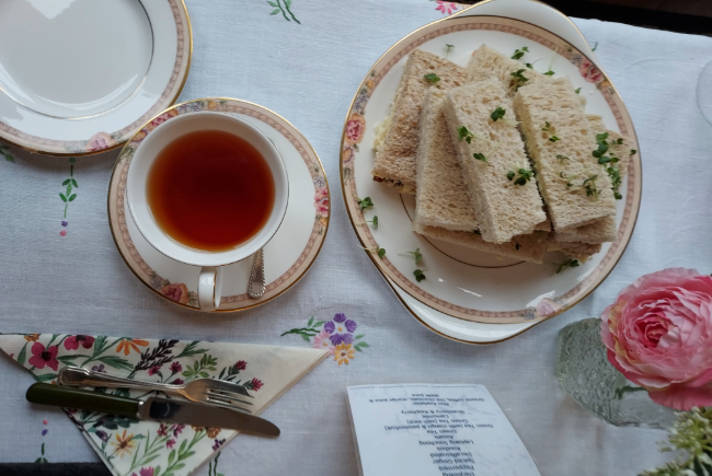 Image shows vintage china on a tablecloth with a cup of tea and plate of sandwiches.